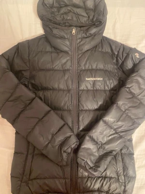 Svart Peak Performance pufferjacka - Svart pufferjacka från Peak Performance med huva och dragkedja framtill. Jackan har quiltad design och logga på bröstet samt ärmen. Tillverkad i syntetmaterial och dunfylld för extra värme. Perfekt för kalla dagar och har en clean, sportig look. (se bild 3,4 för defekter)