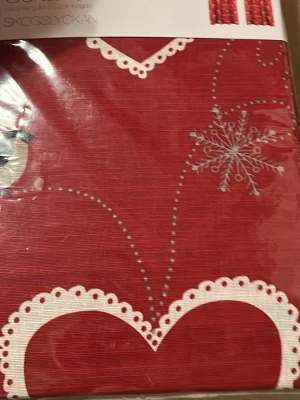 Gardiner  - Säljer en röd väst från SKOGSLYCKAN i 100% bomull med vita spetsdetaljer och snöflingemönster. Västen har dekorativa prickar och hjärtformade kanter som ger en mysig vinterkänsla. Perfekt för dig som gillar juliga och unika plagg.