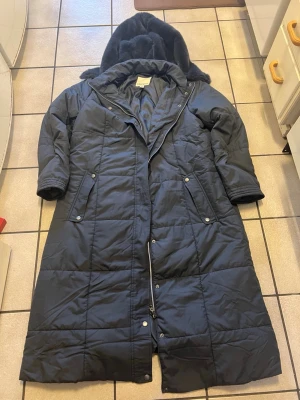 Mörkgrå lång pufferjacka från Bonprix - Lång svart pufferjacka från Bonprix med huva och quiltad design. Jackan har dragkedja och tryckknappar framtill, samt två snedställda fickor med knappstängning. Perfekt för kalla dagar med sin rejäla längd och fodrade insida.