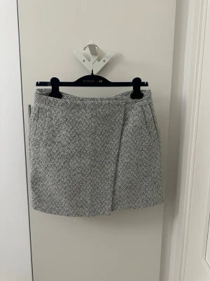 Gråvit rutig kortkjol från New Look - Säljer en snygg kortkjol från New Look i grått och vitt med rutigt mönster. Kjolen har en omlott-look framtill och är tillverkad i ett tjockare, stickat material som känns mjukt. Perfekt för dig som gillar stilrena detaljer och vill ha något som sticker ut.