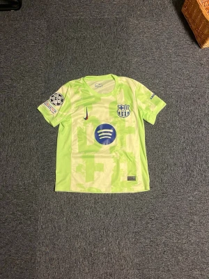 Barcelona fotbollströja  - Bilderna visar en fotbollströja från FC Barcelona med Lamine Yamal och nummer 19, inte några skor. Tröjan är ljusgrön med blå detaljer och UEFA Foundation-märke på ärmen.