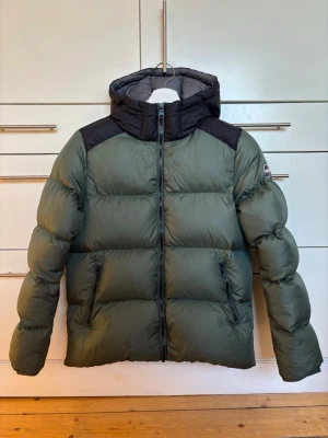 Grön och svart  dunjacka från Colmar storlek 14 år/166 cm/XXS - Grön dunjacka från Colmar med svarta detaljer på axlar och huva. Jackan har en vadderad huva, två dragkedjeförsedda fickor + innerficka och Colmar-logga på ärmen + nederkant rygg. Storlek 14 år/166 cm/XXS. Perfekt skick! Köpt för 4000 på Kid’s Brandstore.