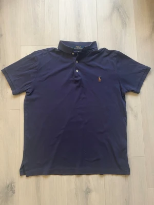 Mörkblå Ralph lauren piké (äkta) - Säljer en mörkblå piké från Ralph Lauren i modellen Custom Slim Fit. T-shirten har korta ärmar, klassisk krage och tre knappar framtill. Broderad Ralph Lauren logga på bröstet. Skick 7/10 inga större synliga defekter men har blivit lite mindre i storleken på grund av tvätten. Storlek S. kontakta mig om du har frågor!