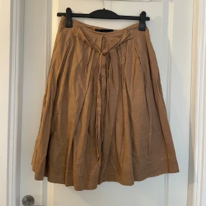 Beige linnekjol från Zara Basic - Superfin beige kjol från Zara Basic i linne med knytband i midjan. Kjolen har en lätt utställd form och fina veck framtill som ger ett snyggt fall. Perfekt för dig som gillar en avslappnad men ändå stilren look.