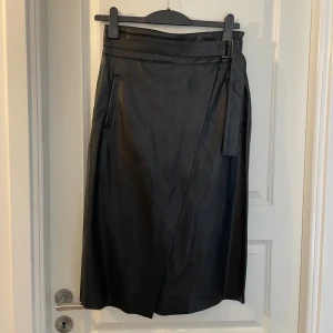 Svart omlottkjol i skinn från Promod - Snygg svart långkjol i skinnimitation från Promod med omlott-design och bälte i midjan. Kjolen har raka linjer, diskreta fickor och dragkedja bak. Perfekt för dig som gillar en stilren och edgy look.