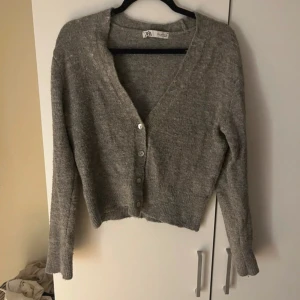 Grå kofta från Zara med knappar - Mysig grå kofta från Zara med v-ringning och fyra knappar framtill. Koftan är stickad och har långa ärmar samt ribbade muddar vid ärmslut och nederkant. Perfekt för lager-på-lager och chill dagar.