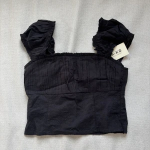 Svart blus med volangärmar från NA-KD - Supersöt svart blus från NA-KD (Moa Mattson kollektion)  med breda volangärmar och broderade detaljer nertill. Toppen har rynkade axelband och diskreta vertikala sömmar upptill.