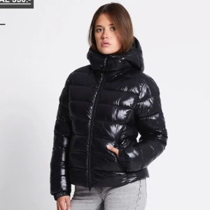 Svart glansig pufferjacka med huva - Säljer en svart, glansig pufferjacka med stor huva och dragkedja framtill. Jackan är vadderad och har en modern, lite boxig passform som håller dig varm. Perfekt för kalla dagar och enkel att styla till jeans eller mjukisbyxor.