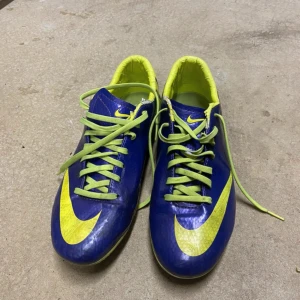 Nike Mercurial blå/gula fotbollsskor - Säljer ett par Nike Mercurial fotbollsskor i blått och gult med limegröna snören och detaljer. Skorna har en smal passform, platt sula med dobbar och är tillverkade i syntetmaterial. Perfekta för dig som vill sticka ut på planen.