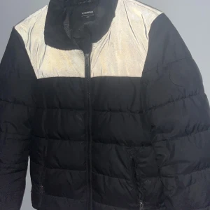 Svart och silver pufferjacka Everest - Säljer en svart pufferjacka från Everest med silvriga detaljer över axlarna. Jackan har hög krage, dragkedja framtill och två fickor med dragkedja. Perfekt för kalla dagar när du vill sticka ut lite extra. Luvan är tyvärr borta