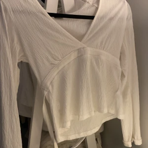 Vit omlottopp från Gina Tricot XS - Säljer en vit omlottopp från Gina Tricot i storlek XS. Toppen har långa ärmar, v-ringning och knytband i midjan. Materialet är lätt strukturerat och ger en snygg och luftig känsla. Perfekt att styla med jeans eller kjol. Helt ny, användt en gång 
