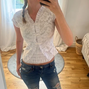Vit spetsblus med volangdetaljer - Supersöt vit blus i spets med blommigt mönster och volangdetaljer längs v-ringningen och framtill. Kortärmad och croppad modell som sitter tajt och har transparent rygg. Inte mina bilder, men bara skriva om fler bilder! Säljer då den är lite stor på mig💕