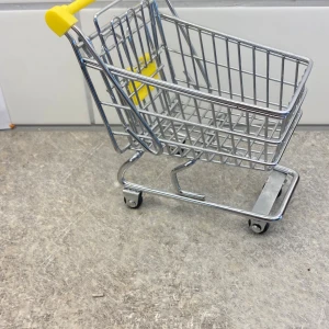 Mini shoppingvagn i metall och plast - En liten shoppingvagn i metall med detaljer i gul plast. Perfekt som dekoration eller för att förvara småsaker. Väskan har klassisk kundvagnsdesign med hjul och handtag i gult. Stilren och rolig accessoar till skrivbordet eller rummet.