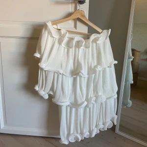 Vit volangklänning från NA-KD - Superfin vit kortklänning från NA-KD med flera lager volanger och plisserat tyg. Klänningen har en lös passform och är ärmlös med offshoulder-stil. 