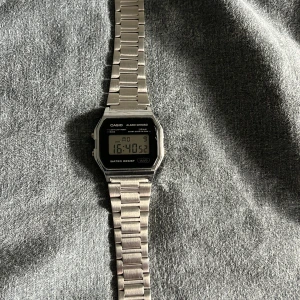 Casio digital klocka i silver - Casio digital klocka med klassisk rektangulär display och metallarmband i silver. Klockan har digital tidvisning, alarm och kronograf-funktion. Snygg retrostil med svart ram runt displayen och tydliga knappar på sidorna. Perfekt för dig som gillar stilren och tidlös design. Liten skråma på själva skärmen