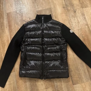 Svart Moncler pufferjacka med stickade ärmar - Snygg svart pufferjacka från Moncler med glansig dunvadderad front och stickade ärmar. Jackan har hög krage, två dragkedjefickor och en dragkedja framtill med röd och vit rand. Perfekt för dig som vill ha en sportig och stilren look. Lappen i nacken har gått av på sidan som man ser i den tredje bilden men det syns inte på och det går att sy 