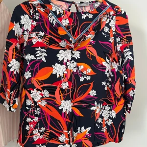 Blommig blus från Fransa i marinblått - Färgstark blus från Fransa med blommigt mönster i vitt, orange och rosa på marinblå botten. Blusen har trekvartsärm, v-ringning med volangdetalj och knapp i nacken. Perfekt för dig som gillar statement-plagg och vill sticka ut.