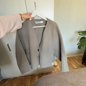 Grå oversized kappa från Zara - Säljer en grå oversized kapa från Zara med v-ringning och två knappar framtill. Jackan har stora fickor och är tillverkad i ett mjukt ull-liknande material. St XS-S