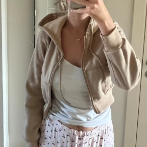 Beige croppad kofta med huva från SHEIN - Mysig beige kofta från SHEIN i storlek XS. Jackan är croppad, har huva, dragkedja framtill och snörning vid halsen. Tillverkad i 100% polyester med ribbade muddar och två fickor framtill. Perfekt för en chill och avslappnad stil.