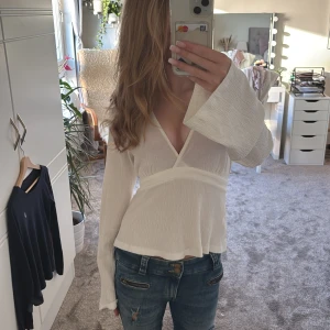 Vit omlottopp med knyt från Gina tricot - Supersnygg vit omlottopp från Bik Bok med v-ringning och långa ärmar. Toppen har knyt i midjan och är gjord i ett lätt, krinklat tyg som ger en chill och luftig känsla. Perfekt till jeans eller kjol för en fräsch look.