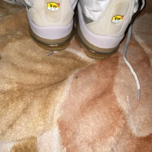 Nike Air Vapormax i ljusgrått - Nike Air Vapormax sneakers i ljusgrått med transparent sula och coola detaljer på ovandelen. Skorna har snörning och en futuristisk design med plastpaneler längs sidorna. Perfekta för dig som gillar streetstyle och vill sticka ut med en modern look.