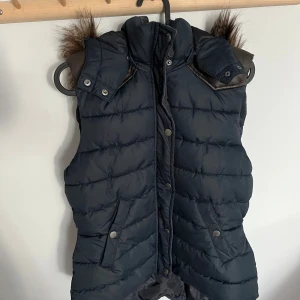 Mörkblå dunväst med fuskpäls från H&M  - Snygg mörkblå dunväst från H&M L.O.G.G. med fuskpäls på luvan och detaljer i mörk brun skinnimitation vid axlarna. Västen har tryckknappar, dragkedja och två fickor framtill. Finns även två spännen (bild 5) på sidan om man vill ha den tajtare. Perfekt för kyliga dagar nu till hösten. Använd fåtal gånger och i mycket bra skick. Storlek 40 men passar även 36/38.
