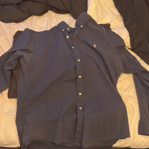 Mörkblå skjorta från Ralph Lauren - Säljer en klassisk mörkblå skjorta från Ralph Lauren med button-down krage och vita knappar. Skjortan har lång ärm och den ikoniska rosa broderade loggan på bröstet. Perfekt för dig som gillar stilrena och tidlösa plagg.