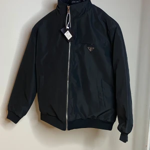 Svart bomberjacka från Prada - Svart bomberjacka från Prada med klassisk siluett och ribbade muddar. Jackan har dragkedja framtill, hög krage och ikonisk Prada-logga i metall på bröstet och ryggen. Tillverkad i slätt syntetmaterial som ger en clean och modern look.