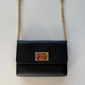 Svart axelväska från Michael Kors - Snygg svart axelväska från Michael Kors i skinn med guldfärgad kedja och detaljer. Väskan har flera fack, kortplatser och en praktisk dragkedja inuti. Perfekt storlek för det viktigaste och stilren design med logoplatta framtill.