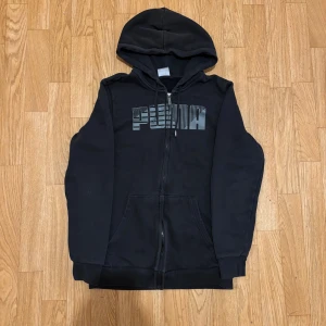 Svart hoodie från Puma med dragkedja - Svart hoodie från Puma med stor logga på bröstet i grått. Tröjan har huva, hel dragkedja och två fickor framtill. Mjuk insida och ribbade muddar vid ärmslut och nederkant. Perfekt för chill eller träning.