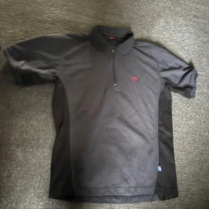 Svart kortärmad t-shirt från Fjällräven - Svart t-shirt från Fjällräven med korta ärmar och krage. Halv dragkedja framtill och rött broderat Fjällräven-logo på bröstet. Materialet känns som bomull eller polyester, perfekt för en chill och sportig look.