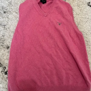 Rosa stickad väst från Gant - Rosa stickad väst från Gant med v-ringning och broderad logga på bröstet. Klassisk modell med ribbade kanter och mjukt material som känns skönt mot huden. Perfekt att slänga över en skjorta för en preppy look.