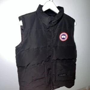 Svart dunväst från Canada Goose - Svart dunväst från Canada Goose med hög krage och klassisk logotyp på bröstet. Västen har stora fickor framtill och stängs med både dragkedja och knappar. Perfekt för lager-på-lager och håller dig varm under kyliga dagar. Storlek XS men passar S också. 