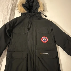 Svart parkajacka från Canada Goose XS - Säljer en svart parkajacka från Canada Goose i storlek XS. Jackan har stor huva med fuskpäls, flera stora fickor framtill och klassisk Canada Goose-logga på bröstet. Yttermaterialet är polyester och bomull, fodrad med dun och har snygga detaljer.