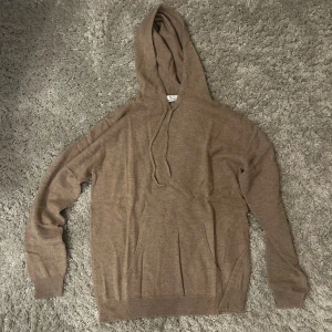 Brun kashmir hoodie med huva - Mysig brun kashmir hoodie med huva och snörning. Tröjan har långa ärmar och ribbade muddar vid ärmslut och nederkant. Perfekt för kyliga dagar när du vill vara både bekväm och snygg.