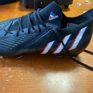 Svarta Adidas fotbollsskor med mönster - Säljer ett par svarta Adidas fotbollsskor med klassiska tre ränder i vitt, rött och blått på sidan. Skorna har snörning, högre strumpliknande skaft och mönstrad ovansida för extra grepp. Sulan har fasta dobbar och skon är tillverkad i syntetmaterial.