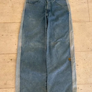 Wide jeans från Anvil med gator-detalj - Säljer ett par ljusblå jeans från Anvil med bred passform och cool gator-patch på bakfickan. Jeansen är tillverkade i 100% bomull och har klassisk femficksdesign. Perfekta för dig som gillar retrovibbar och unika detaljer.