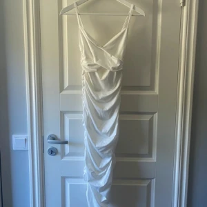 Vit omlottklänning från SHEIN - Säljer en lång vit klänning från SHEIN i storlek XS. Klänningen har smala axelband, omlott upptill och snygg rynkad effekt längs hela framsidan. Materialet är mjukt och har en lätt satinliknande känsla. Perfekt för dig som vill ha en elegant och trendig look.