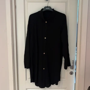 Svart långärmad skjorta  - Svart långärmad skjorta med klassisk krage och knäppning framtill. Skjortan har en loose passform och är tillverkad i ett lätt och mjukt tyg, perfekt för en avslappnad stil. Enkel design utan mönster eller extra detaljer.