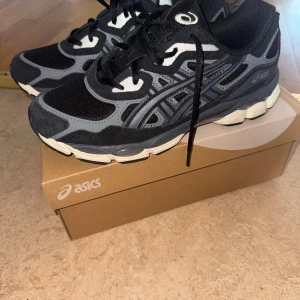 Svarta och grå Asics sneakers - Snygga Asics sneakers i svart och grått med vita detaljer. Skorna har mesh- och syntetpaneler för bra andningsförmåga och komfort. Klassisk snörning och rund tå, samt en vit platt sula som ger en sportig look. Perfekta för dig som gillar streetwear och sportig stil.