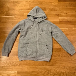 Grå hoodie från Polo Ralph Lauren - Grå hoodie från Polo Ralph Lauren med dragkedja och huva. Klassisk design med snörning i huvan, två fickor fram och broderad logga på bröstet. Perfekt för chill dagar eller när du vill ha en enkel och stilren look.