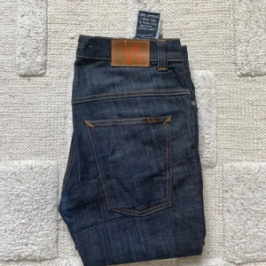 Mörkblå jeans från Nudie Jeans - Säljer ett par mörkblå jeans från Nudie Jeans med klassisk femficksdesign och kontrastsömmar i orange. Jeansen har rak passform och är tillverkade i kraftig bomullsdenim. Snygg läderpatch med logga baktill i midjan. Perfekta för dig som gillar stilrena och tidlösa jeans.