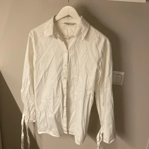 Vit skjorta med öppen rygg från NA-KD - Snygg vit skjorta i bomull från NA-KD x Angelica Blick. Skjortan har långa ärmar med knytband vid ärmsluten, klassisk krage och knappar framtill. Baksidan har en cool öppen rygg-detalj med knytband. 