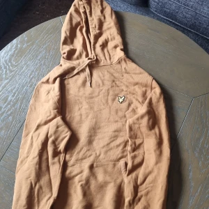 Brun hoodie från Lyle & Scott - Säljer en snygg brun hoodie från Lyle & Scott med klassisk logga på bröstet. Tröjan har huva med snörning och en stor magficka. Perfekt för dig som gillar en avslappnad och stilren look.