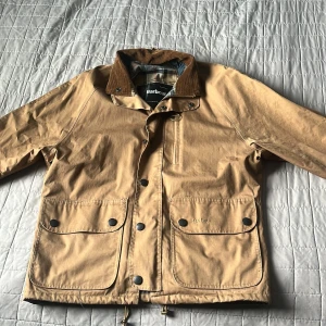 Beige Barbour jacka med detaljer - Snygg beige vindjacka från Barbour med klassisk krage i manchester, stora fickor med tryckknappar och dragkedja framtill. Jackan har rutigt innerfoder och coola detaljer som metallringar och broderad logga på fickan. Perfekt för dig som gillar stilren och tidlös look.