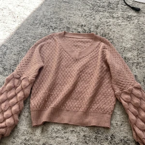 Rosa stickad tröja med puffärmar - Superfin stickad tröja i dammigt rosa med v-ringning och unika puffärmar med flätstickat mönster. Tröjan har ribbade muddar och en croppad passform som ger en trendig look. Perfekt för lager-på-lager under kyliga dagar.