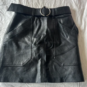 Svart skinnkjol med bälte och fickor - Snygg svart skinnkjol med bredt bälte och silverfärgat spänne. Kjolen har två stora fickor framtill och markerade sömmar som ger en cool look. Perfekt för dig som vill ha en edgy stil och gillar detaljer som sticker ut.