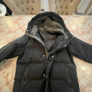 Svart pufferjacka EA7 Emporio Armani - Svart pufferjacka från EA7 Emporio Armani med huva och dragkedja. Jackan har logga på bröstet och stor Emporio Armani-tryck på ryggen. Snygga reflexdetaljer längs ärmarna och två dragkedjeförsedda fickor framtill. Perfekt för kalla dagar. Storlek L men sitter som en vanlig M