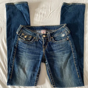 True Religion jeans lågmidjade - Blå jeans från True Religion med tydliga kontrastsömmar och ikoniska bakfickor med guldfärgade knappar. Låg midja och raka ben. De har små slitningar vid hälarna men inte något som är märkbart, annars fint skick. Strl 26, säljer eftersom de var för stora. Fraktsr gärna tillsammans med andra plagg.