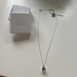 Silverhalsband med blå droppe från Caroline Svedbom - Stilrent halsband i silver från Caroline Svedbom med en droppformad blå kristall som hängsmycke. Kedjan är tunn och elegant, perfekt för att ge en lyxig touch till din outfit. Levereras i original ask. Säljs då halsbandet ej kommer till användning då jag har guld och inte sover smycken. Därav att halsbandet är oanvänt :)    Nytt pris på halsbandet är 800:- 